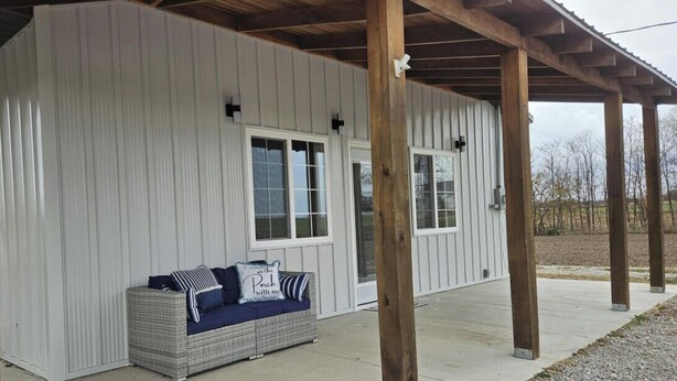 A covered porch with a wicker sofa and cushions, a white exterior wall, and wooden beams supporting the roof.