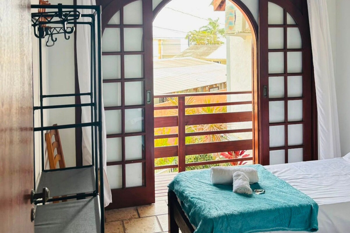 A room with a bed, a ceiling fan, a window with a view, and a door with a metal gate.