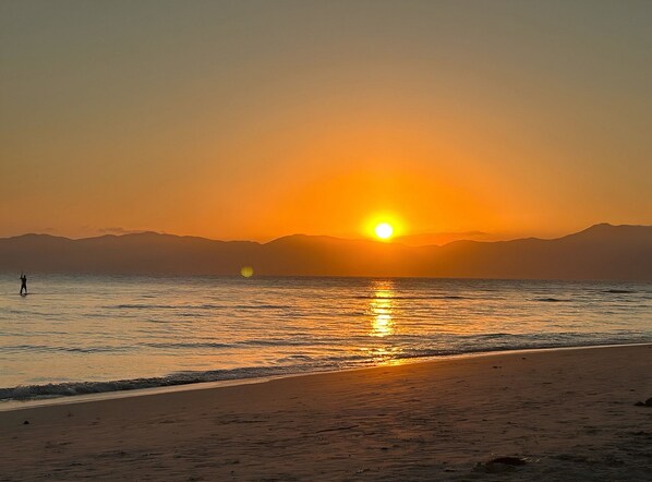 A beach with a sunset, calm sea, and distant mountains.