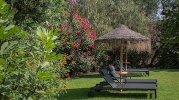 Een tuin met een rieten parasol, twee loungestoelen en diverse bloeiende planten.