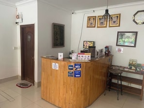 A reception desk with a wooden finish, a small stool, and a bench. There are signs for ABA and VISA, a framed picture, and a wall-mounted clock. - Kaya Hotel  (Krong Siem Reap)