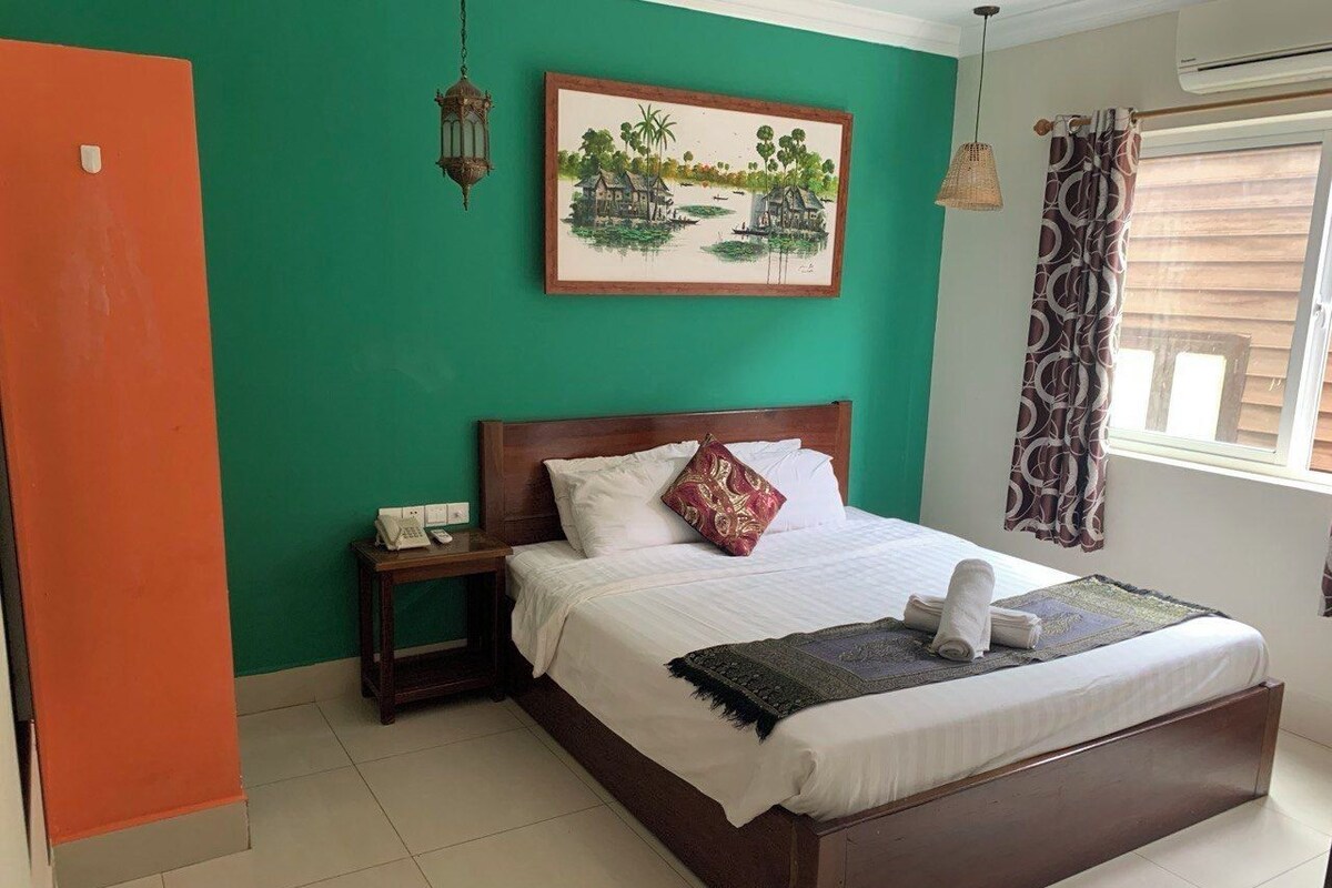 A hotel room with a bed, a nightstand, a telephone, a painting, and a window with curtains.