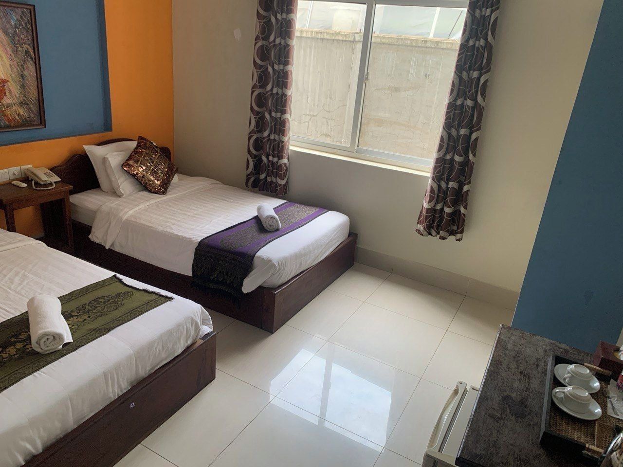A hotel room with two beds, a bedside table with a phone, a window with curtains, and a small kitchenette area.