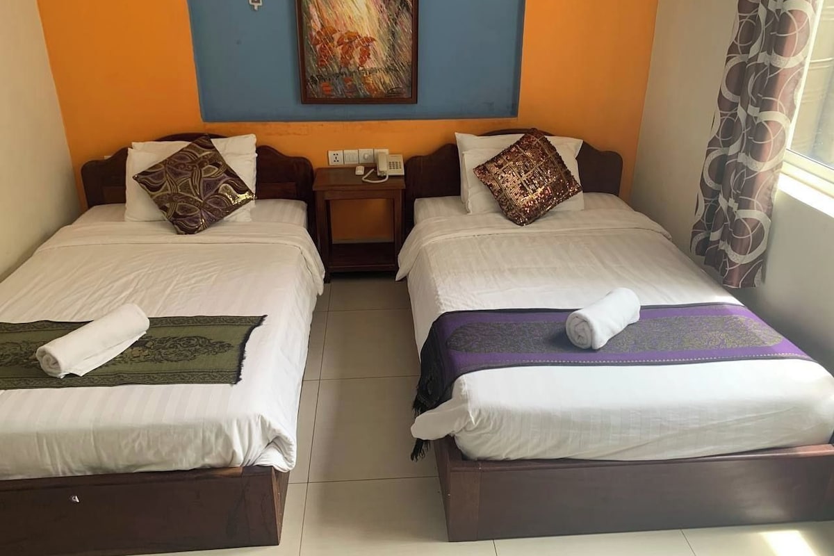 A hotel room with two single beds, a nightstand, a painting on the wall, and a window with curtains.