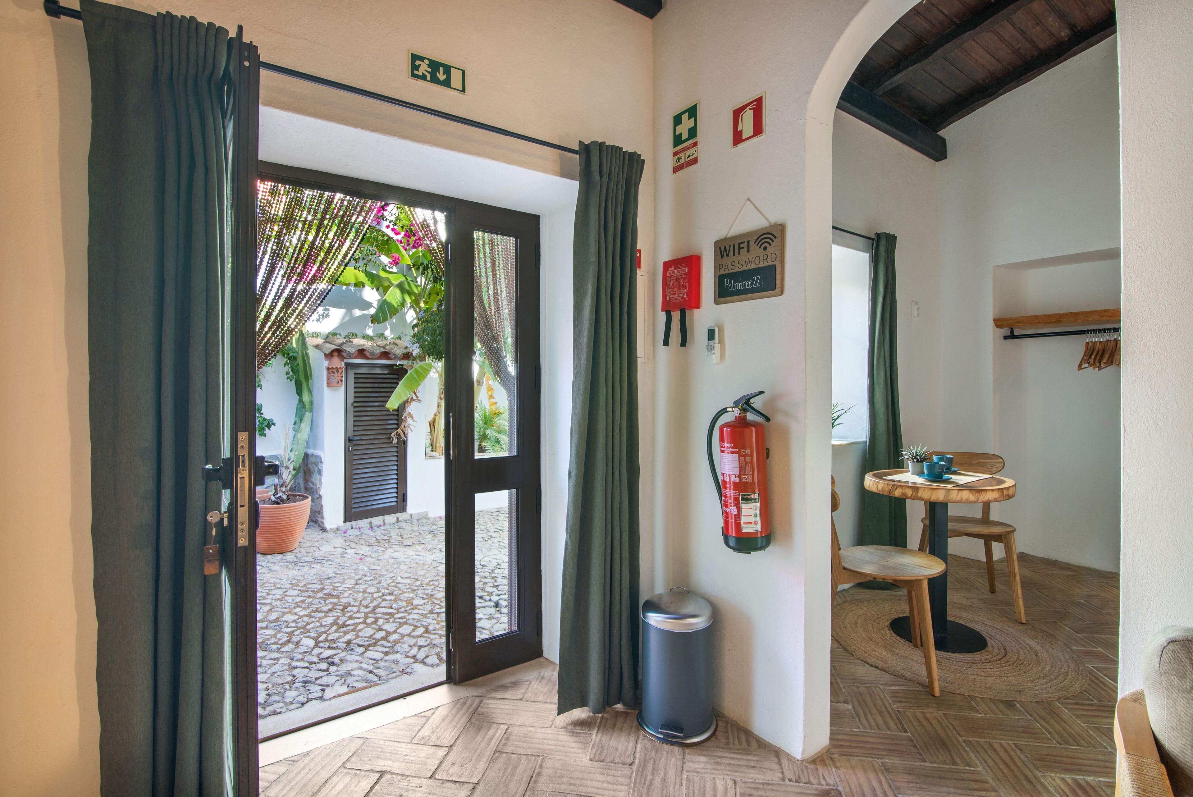 Quarto com piso de madeira, uma pequena mesa com cadeiras, um extintor de incêndio e uma porta que dá para o exterior.