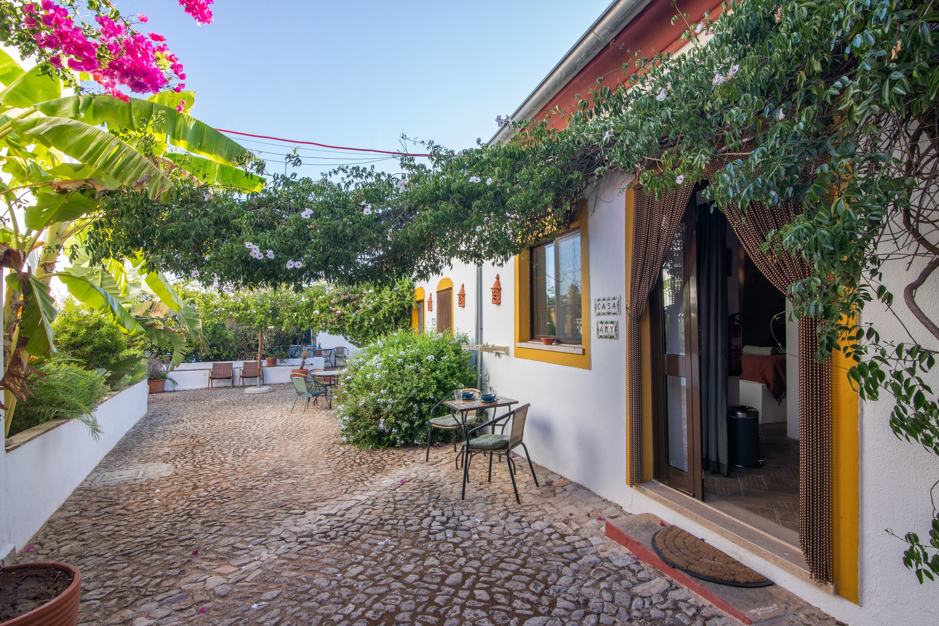 Un patio con suelo de adoquines, una mesa con sillas y un edificio con un ventanal amarillo y un letrero que dice 'CASA'.