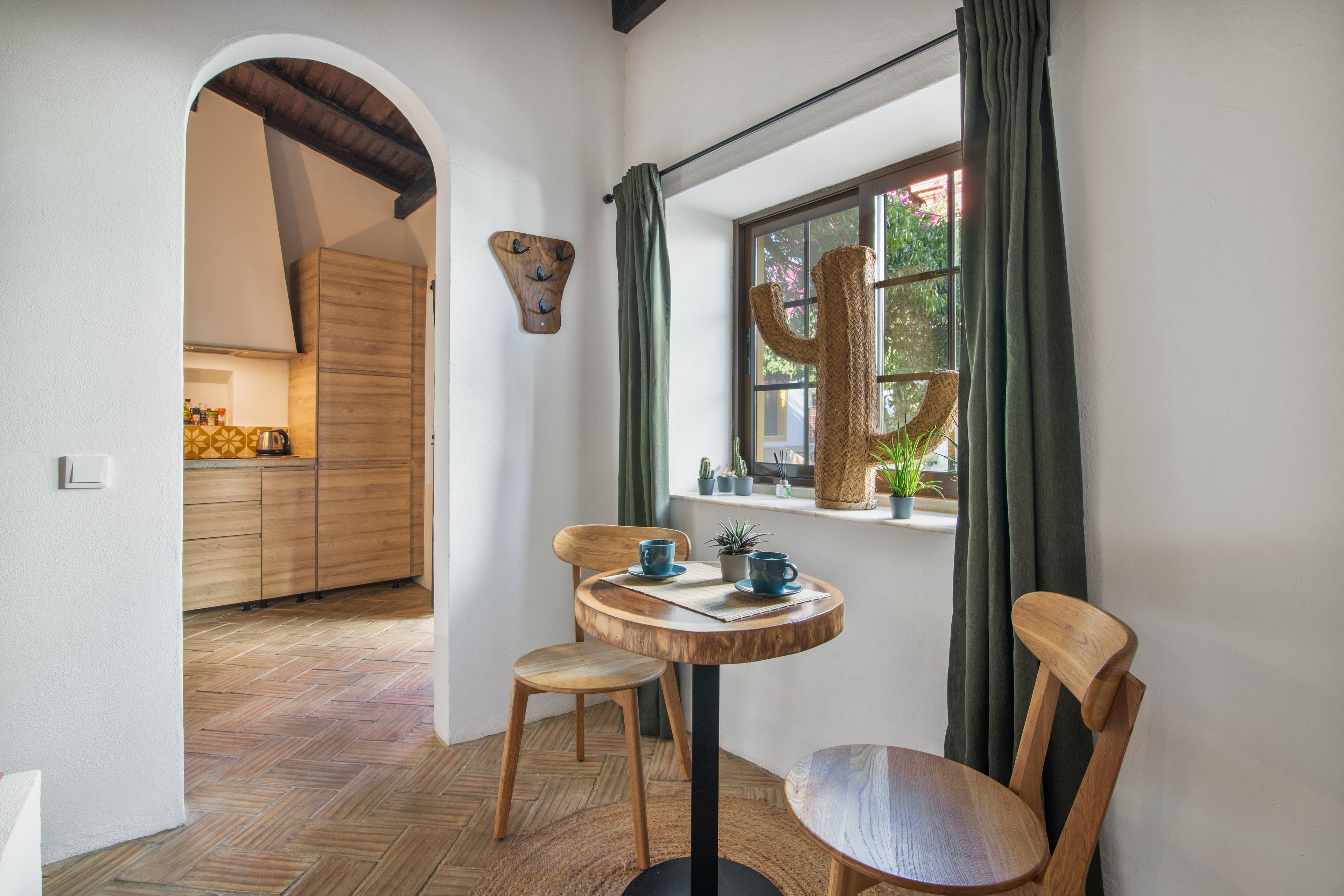 Quarto com mesa de jantar de madeira, duas cadeiras, janela com vista para áreas verdes e cozinha ao fundo.
