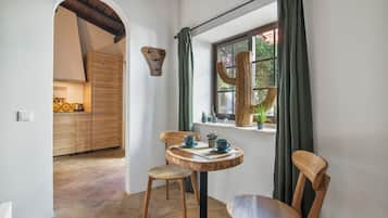 Quarto com mesa de jantar de madeira, duas cadeiras, janela com vista para áreas verdes e cozinha ao fundo.