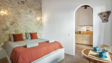 Une chambre avec un mur en pierre, un lit avec une literie orange et une vue sur la cuisine par le biais d’une arche.