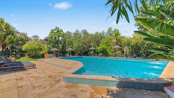 Una piscina con borde de azulejos, rodeada de sillones de descanso y sombrillas, ubicada en un jardín ajardinado con diversas plantas y árboles.