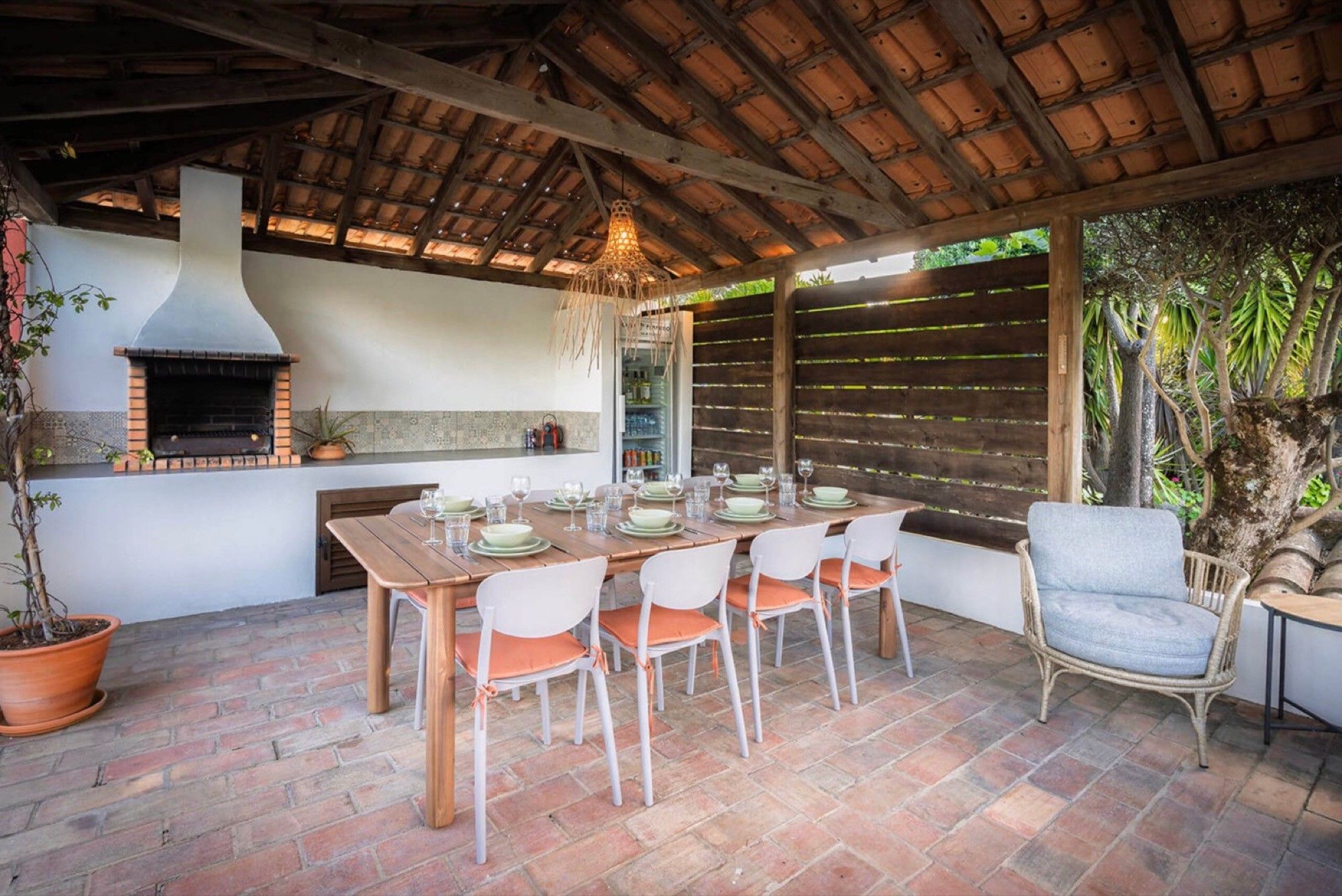 An outdoor dining area with a brick floor, wooden table, white chairs, and a brick oven.