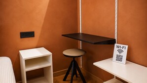 Una habitación pequeña con paredes anaranjadas, un escritorio blanco, un taburete y una estantería. Hay un cartel que indica acceso a Wi-Fi.