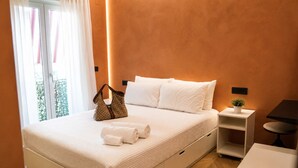 Una cama bien hecha con ropa blanca, un bolso marrón y toallas enrolladas sobre un piso de madera.