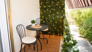 Un pequeño patio al aire libre con una mesa redonda y dos sillas, una planta en maceta y una pared cubierta de follaje y luces colgantes.
