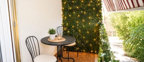 A small outdoor patio with a round table and two chairs, a potted plant, and a wall covered in greenery and string lights.