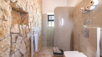 A bathroom with a stone wall, a white toilet, a shower area, and a basket with towels.
