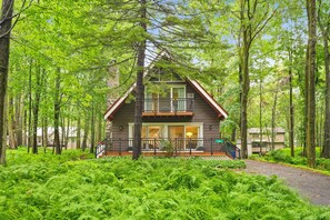 A two-story cabin with a balcony, surrounded by dense green foliage. - A-Frame Retreat - Hot Tub, Fire Pit & Fireplace (Monroe County)