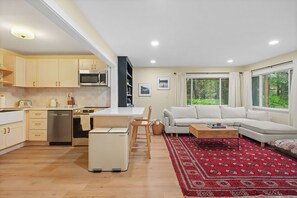 A modern kitchen with a microwave, stove, and cabinets, a dining area with a table and chairs, a living room with a sofa and a red patterned rug, and large windows with a view of greenery. - A-Frame Retreat - Hot Tub, Fire Pit & Fireplace (Monroe County)