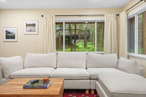 A modern living room with a sectional sofa, a coffee table with a book and remote, and a large window with curtains. - A-Frame Retreat - Hot Tub, Fire Pit & Fireplace (Monroe County)