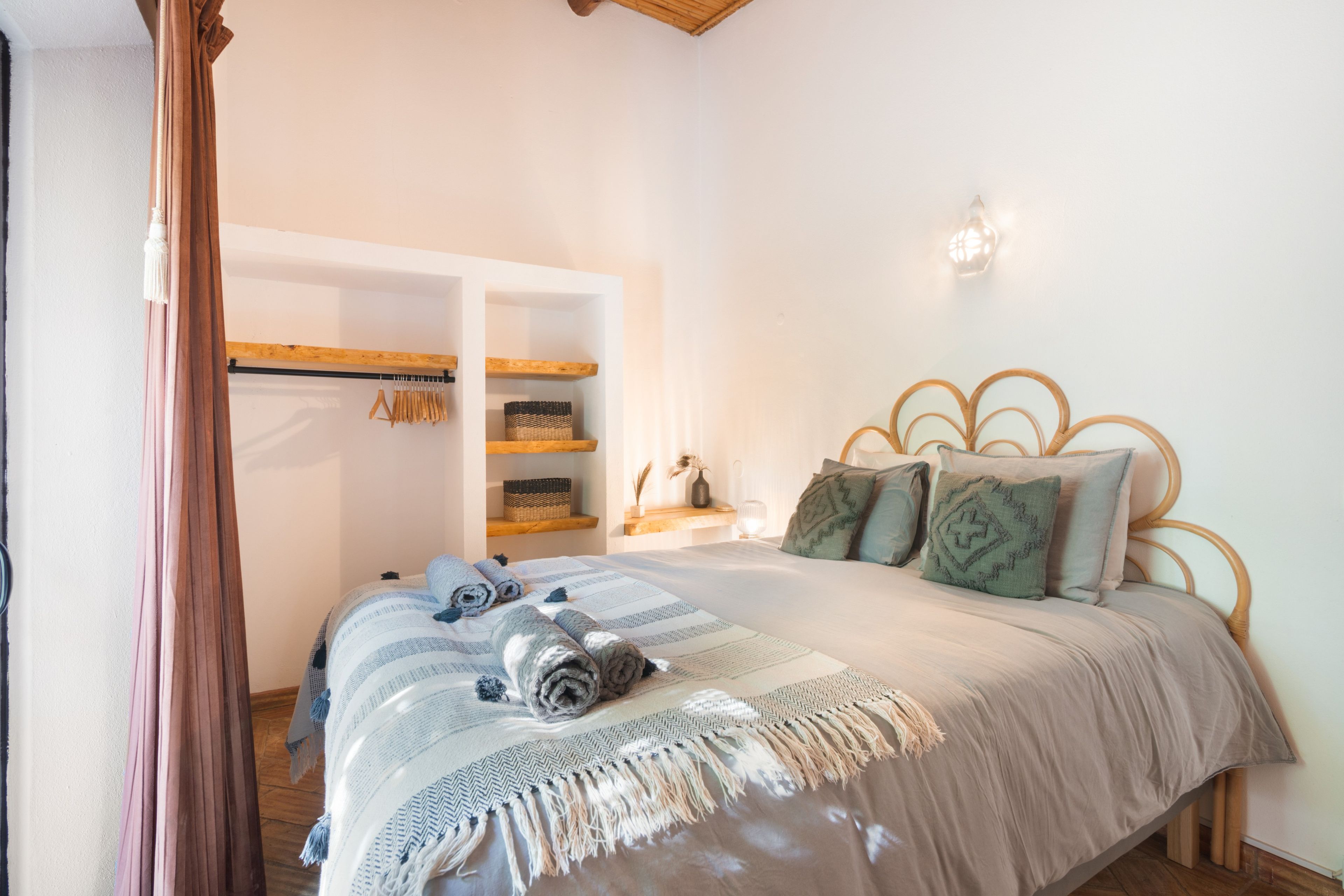 Une chambre à coucher comprenant un lit, une tête de lit en bois, une table de chevet, une lampe fixée au mur et une armoire avec des vêtements suspendus.