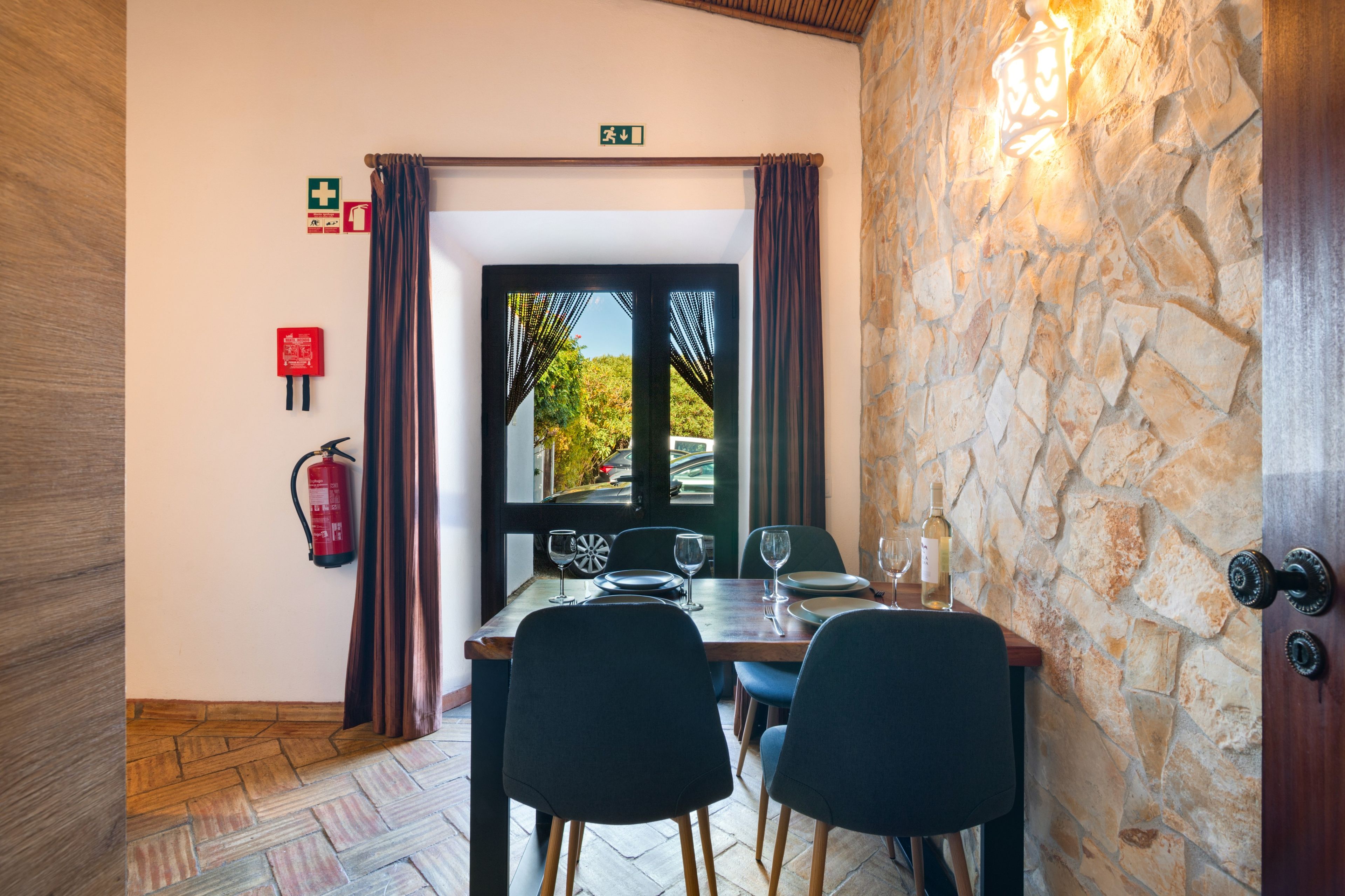 A dining area with a stone wall, a fire extinguisher, and a view through a door.