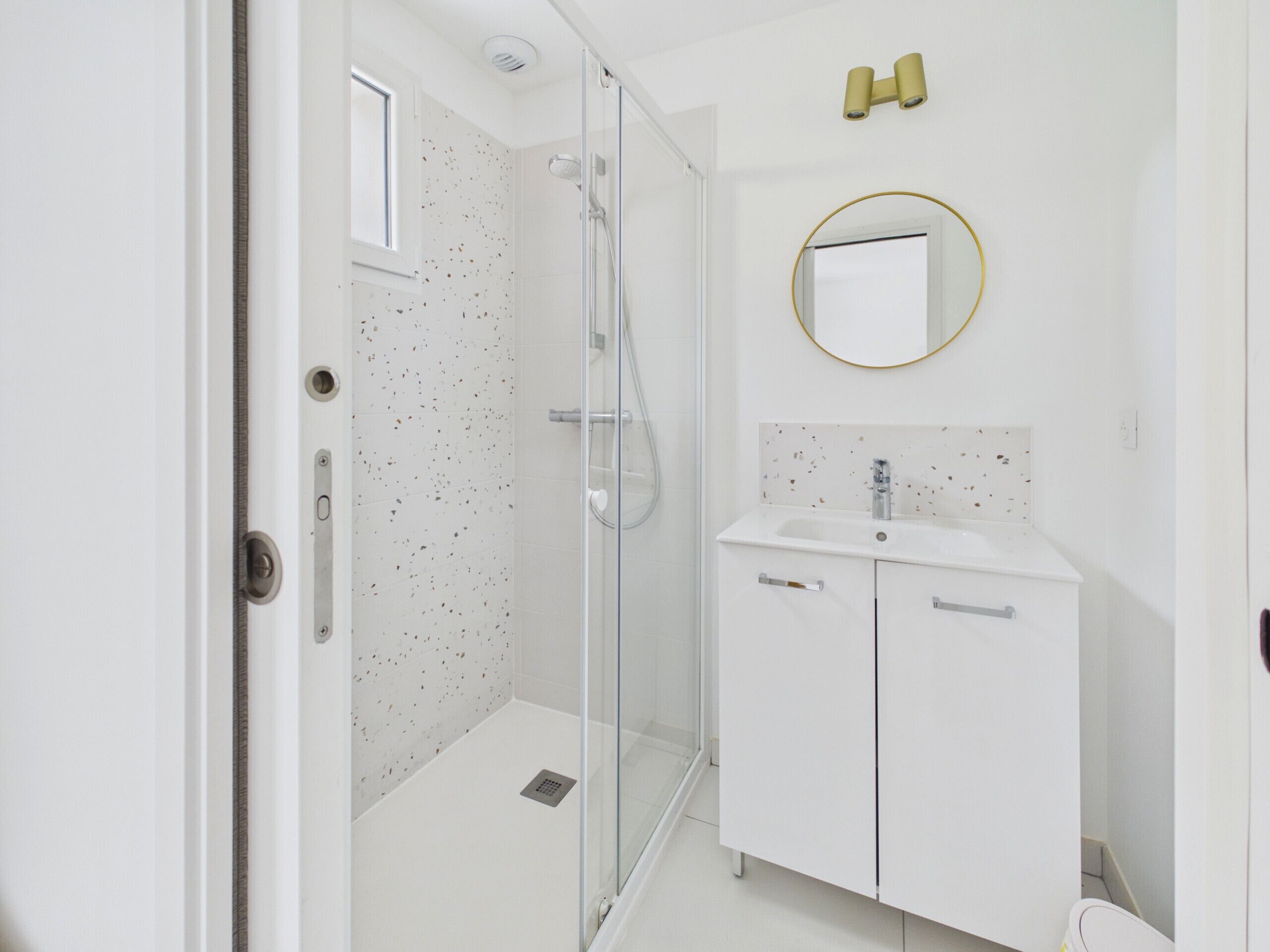 Une salle de bain moderne avec une douche à l’italienne en verre, un lavabo blanc agrémenté d’un miroir doré, et un mur recouvert de carreaux blancs à motifs.