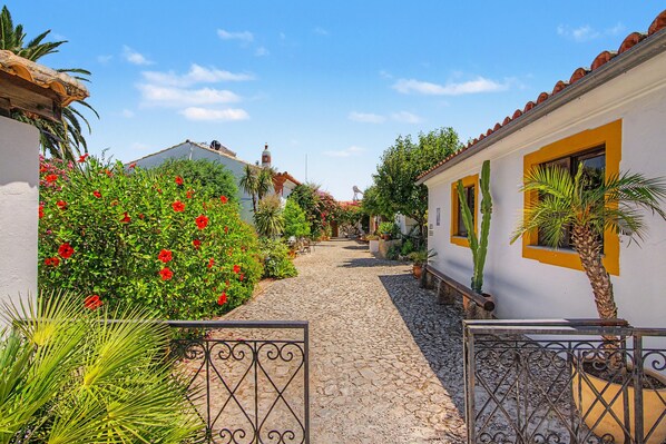 A cobblestone pathway leading through a courtyard with white buildings, red flowers, and green plants.