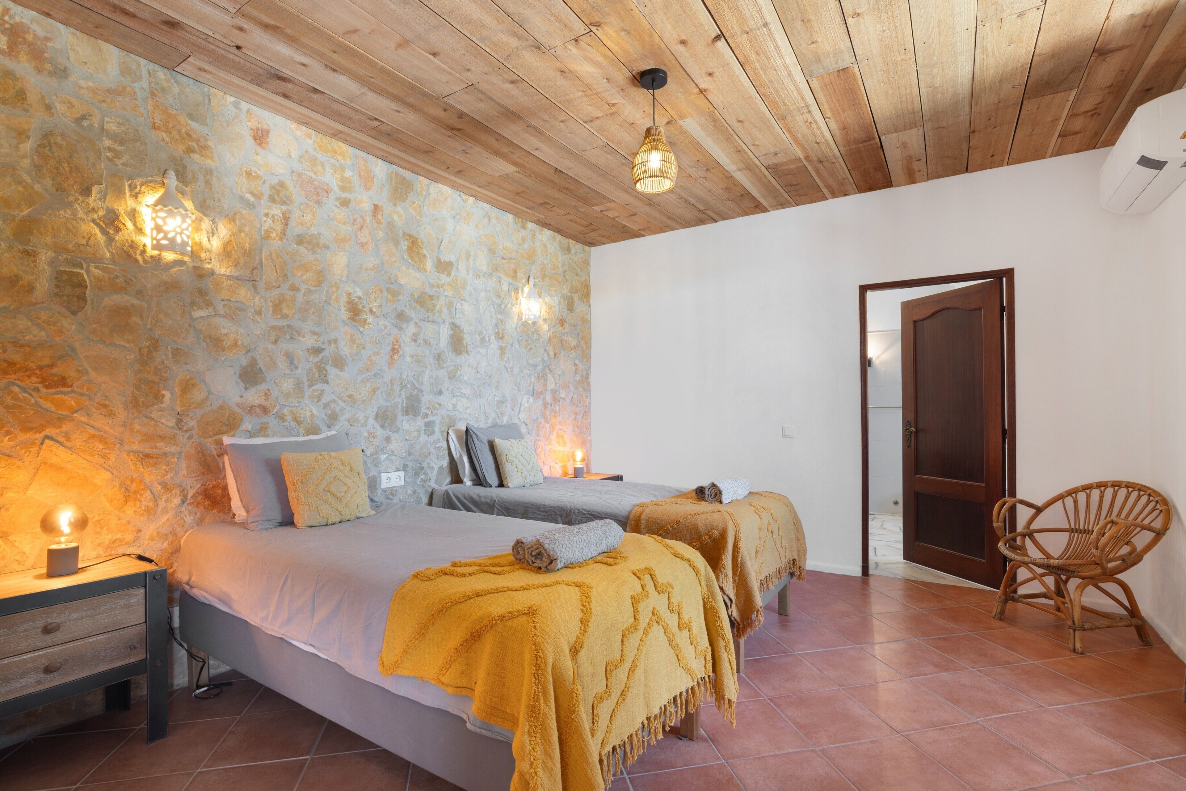 Une chambre avec un mur en pierre, un plafond en bois, un lit avec des oreillers, une table de chevet, une chaise et une porte.