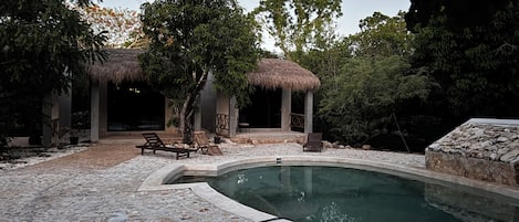 A pool area with a stone deck, lounge chairs, and a thatched-roof structure.