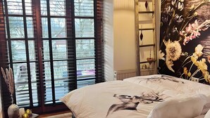 Een slaapkamer met een bed, twee hanglampen, een raam met zonwering, en een wand met een bloemmotief.