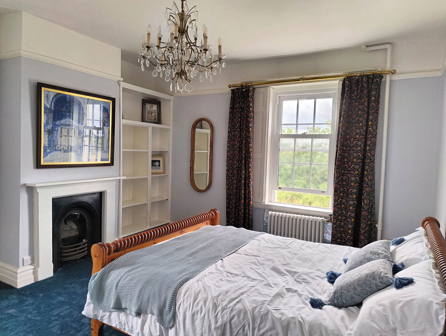 A bedroom with a bed, a fireplace, a chandelier, a window with curtains, and a painting on the wall.