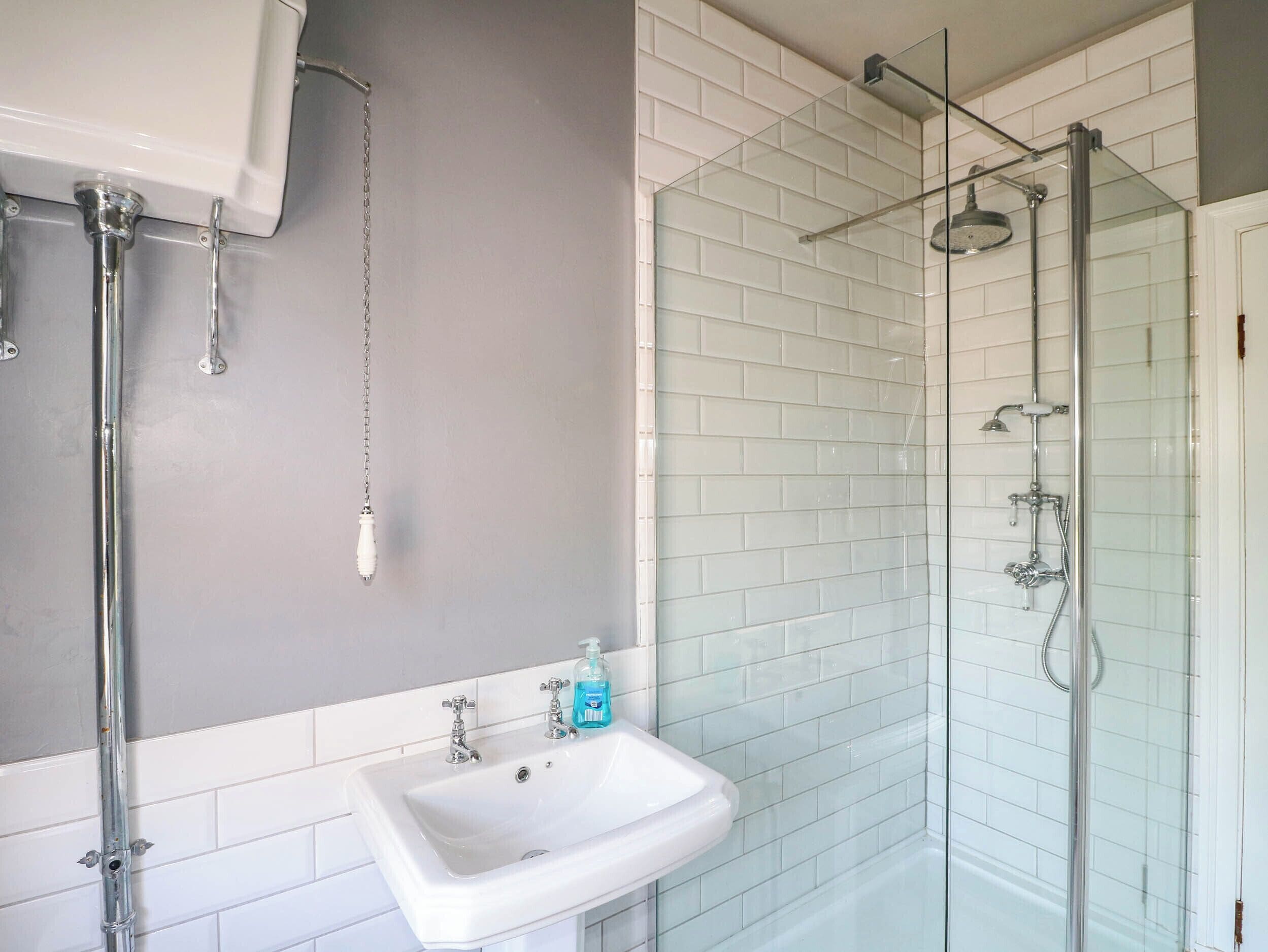 Un baño con lavamanos blanco, cabina de ducha de vidrio y pared de azulejos blancos.
