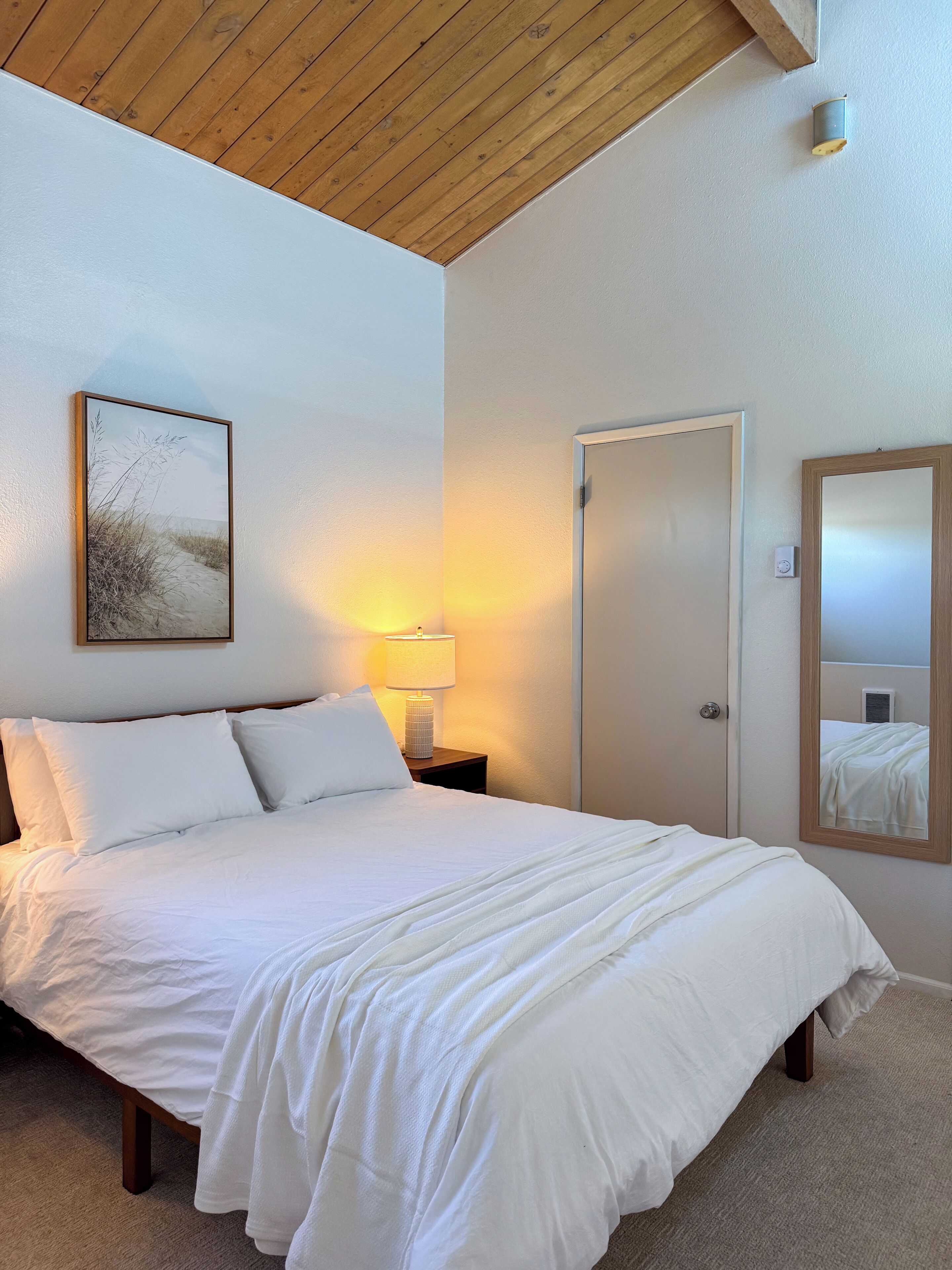 A hotel room with a bed, a bedside lamp, a framed picture on the wall, a door, and a mirror.