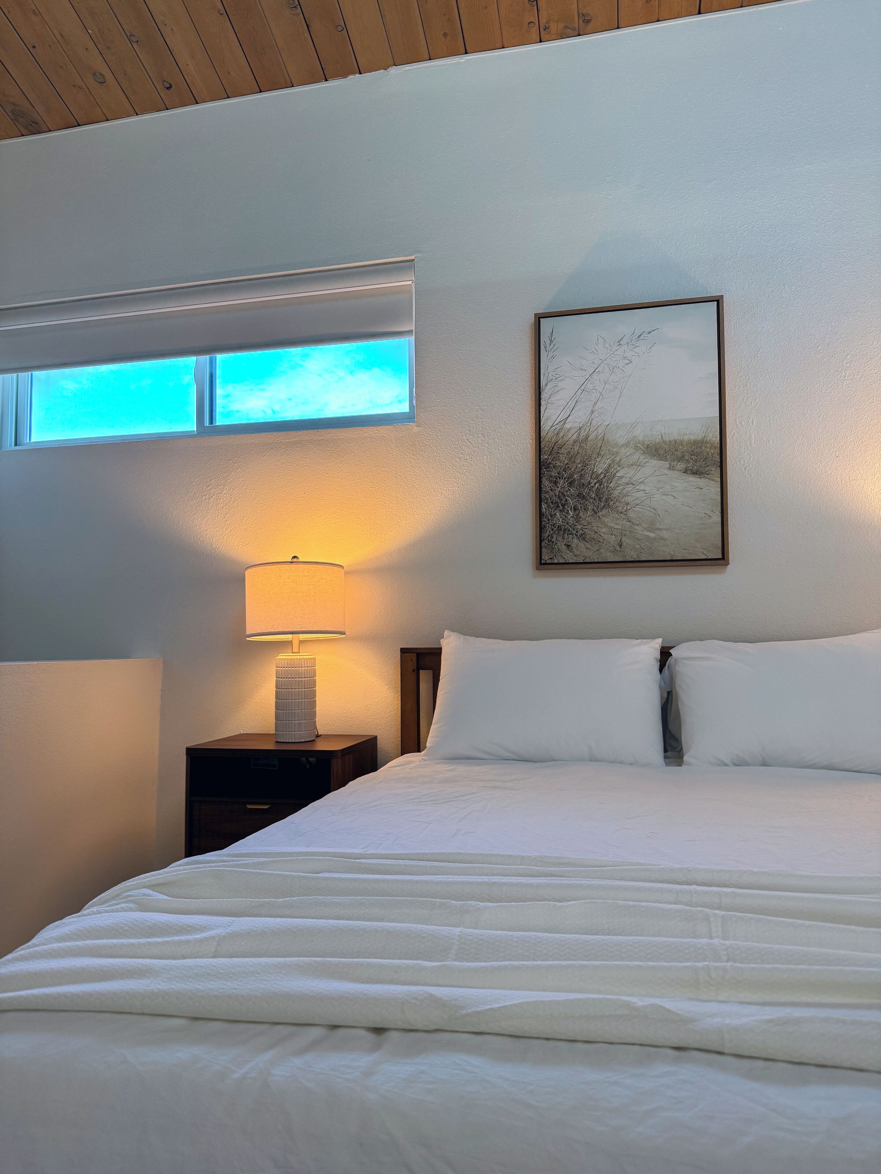 Habitación de hotel con cama, mesita de noche, lámpara, ventana y un cuadro colgado en la pared.