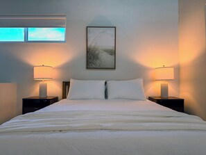 A neatly made bed with white linens, two bedside lamps, a framed picture on the wall, and a window with blinds.