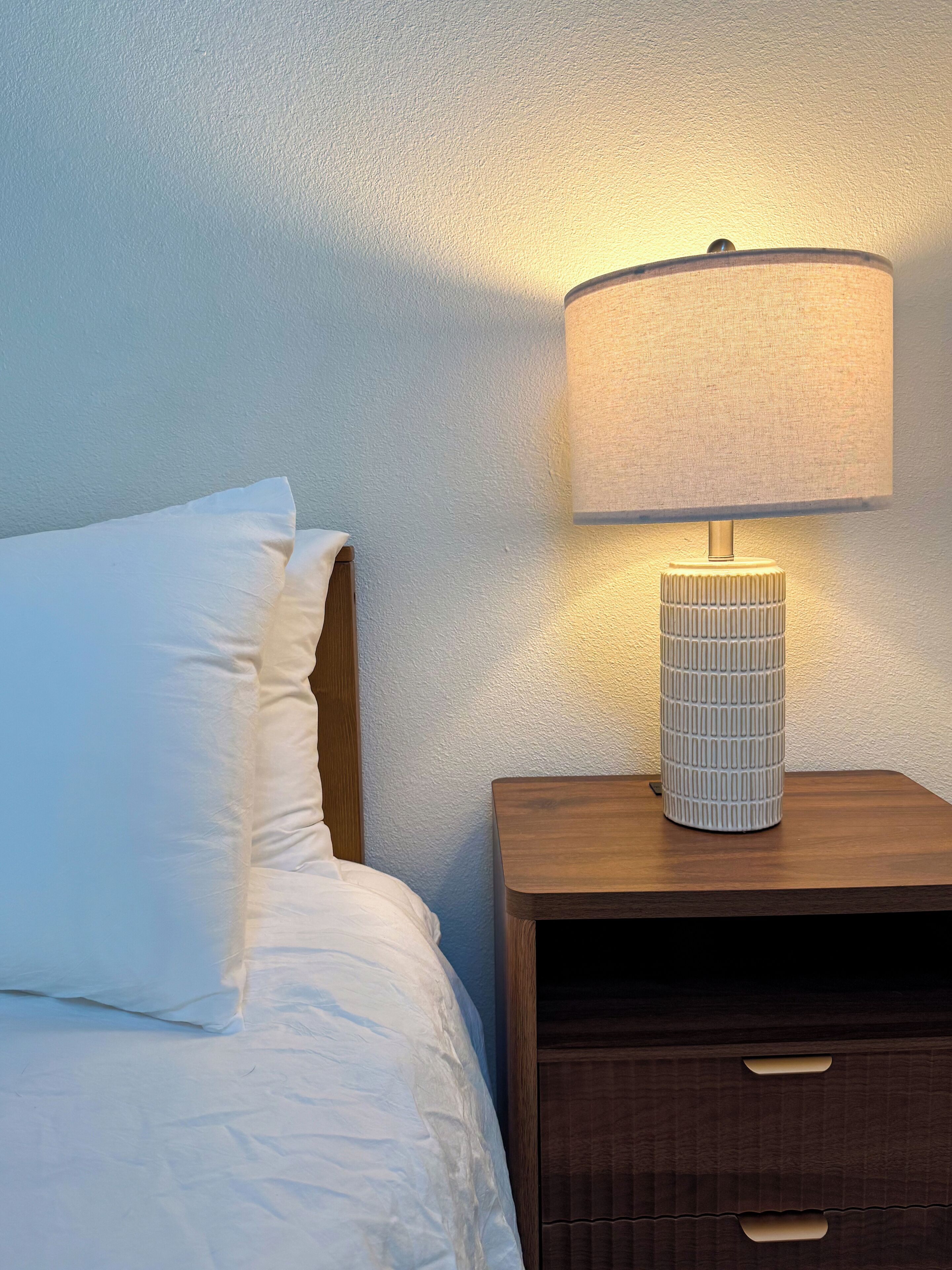 A neatly made bed with white linens, a wooden nightstand with a lamp, and a textured wall.
