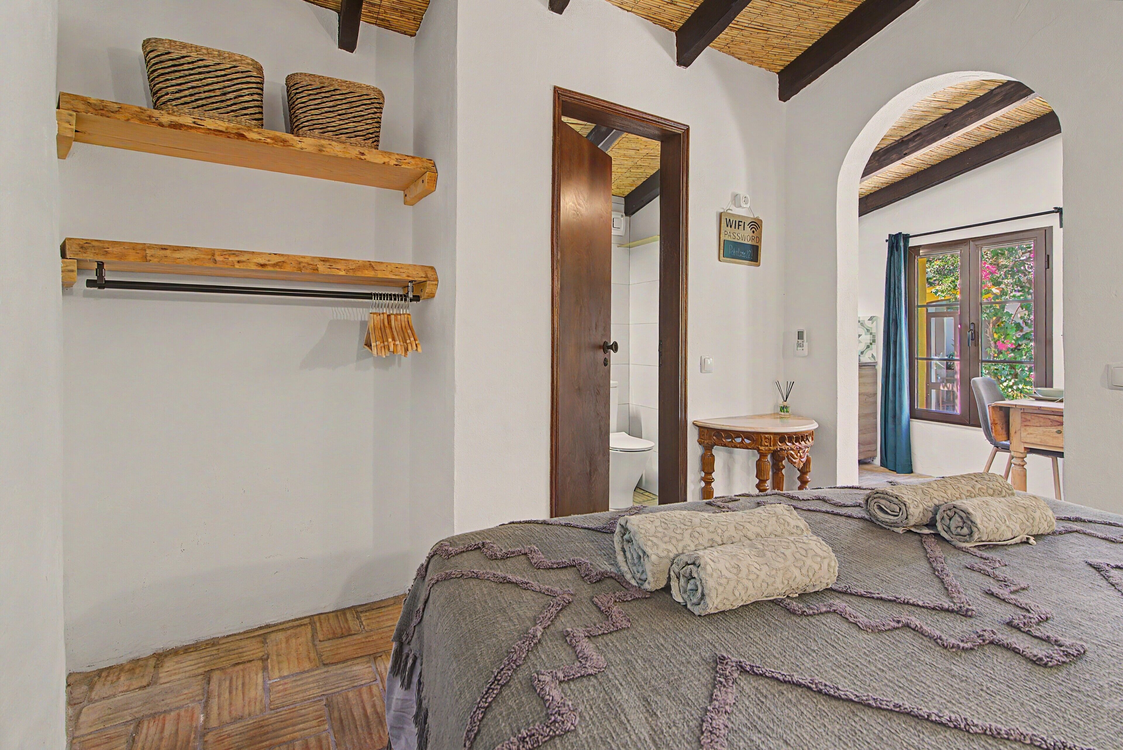 Une chambre à coucher avec une tête de lit en bois, un lit recouvert d’une couverture à motifs, une étagère en bois avec des paniers, une porte en bois et une vue sur un jardin par la fenêtre.