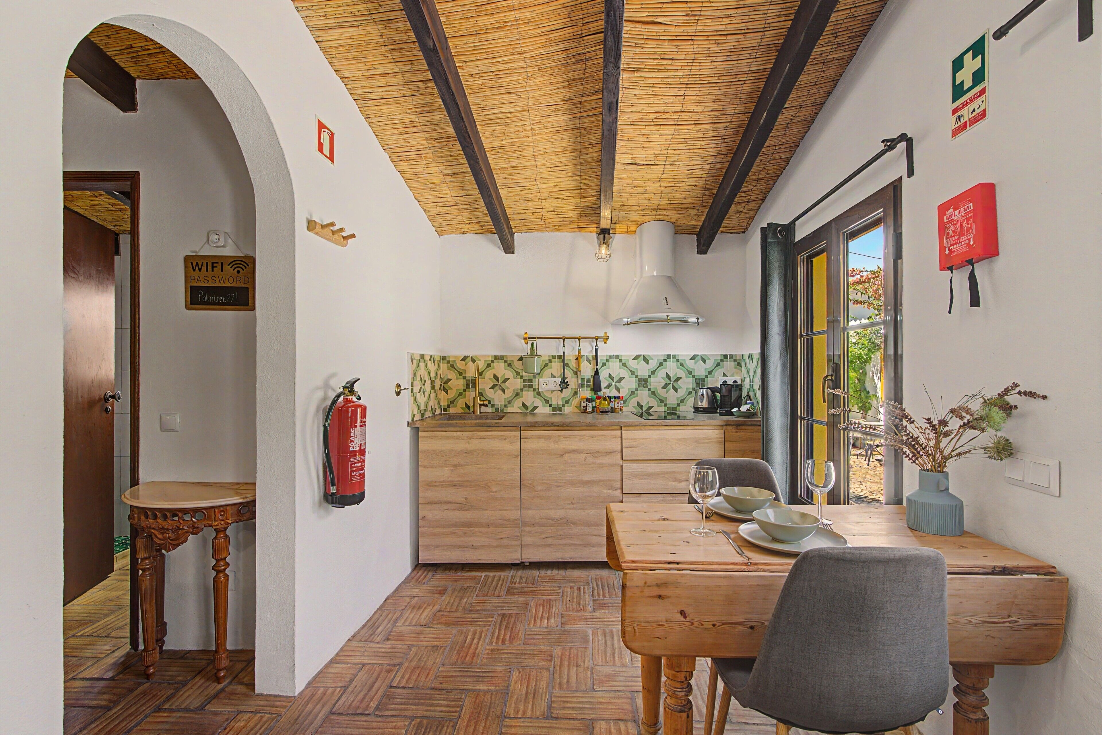 A dining area with a wooden table, chairs, a fire extinguisher, and a kitchen in the background.