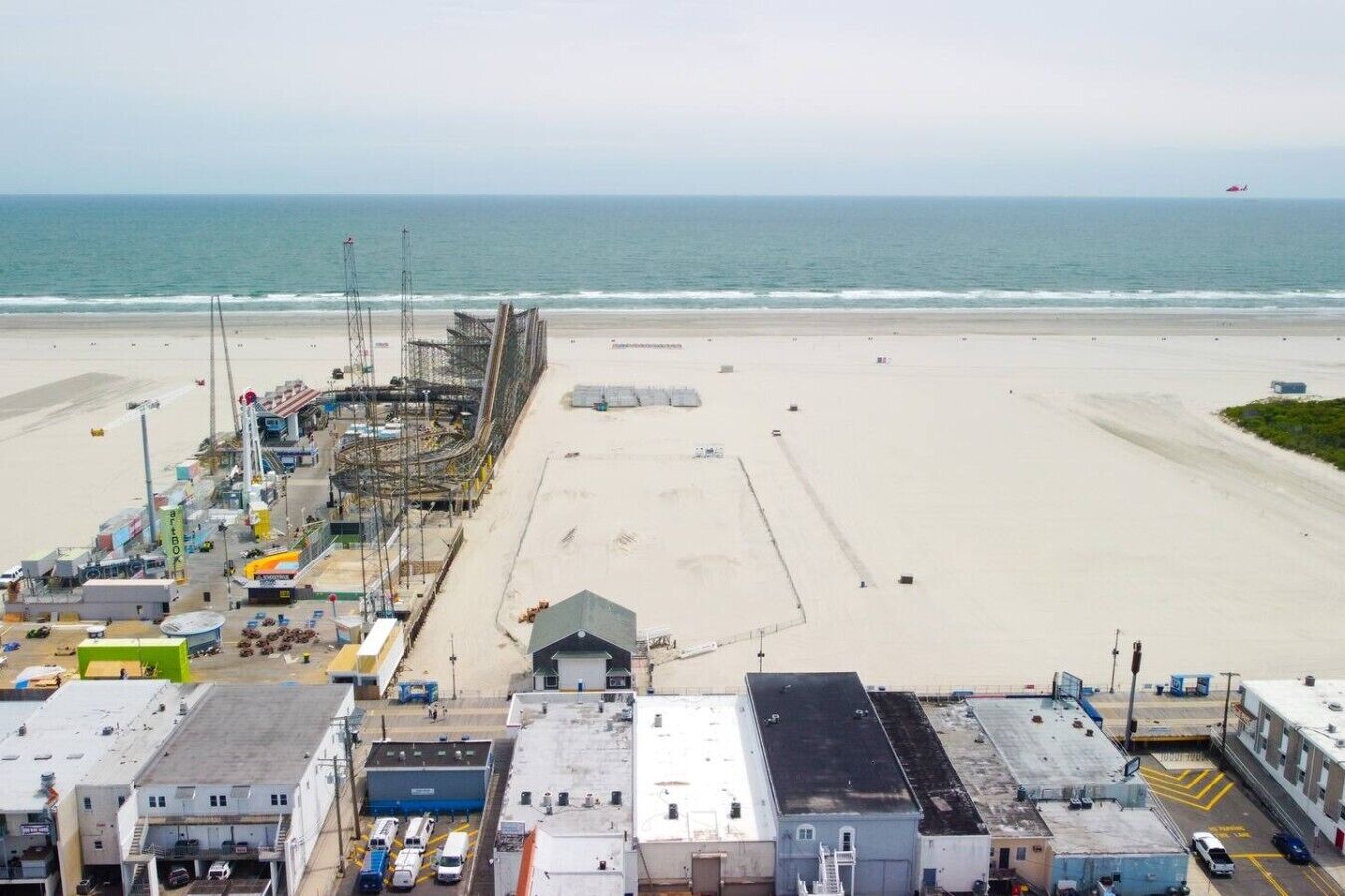 Vista frente al mar con una playa arenosa, un muelle con maquinaria de construcción y un edificio de techo plano en primer plano.