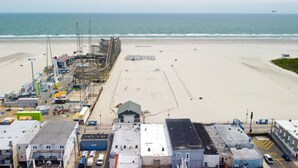 Vista da praia com areia dourada, um cais e equipamentos de construção, e um edifício com telhado plano ao primeiro plano.