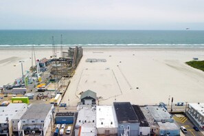 Blick auf einen Strand mit sandigem Ufer, einem Steg mit Baugeräten und einem Gebäude mit flachem Dach im Vordergrund.