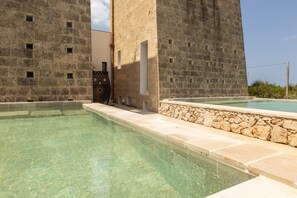 Ein Schwimmbecken mit klarem Wasser, umgeben von Steinmauern und einem modernen Gebäude mit mehreren Fenstern.