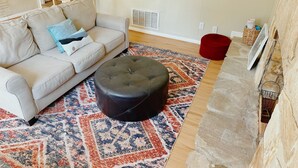 Una sala de estar con un sofá color beige, un reposapiés negro, un tapete estampado, un taburete rojo y una chimenea de piedra.