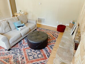 Ein Wohnzimmer mit einem beigen Sofa, einem schwarzen Hocker, einem gemusterten Teppich, einem roten Schemel und einem Kamin aus Stein.
