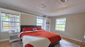 Un dormitorio con una cama, dos ventanas, una mesita de noche y una estantería.