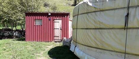 A red shed with a door and a window, a covered trailer, and a grassy area with trees in the background.