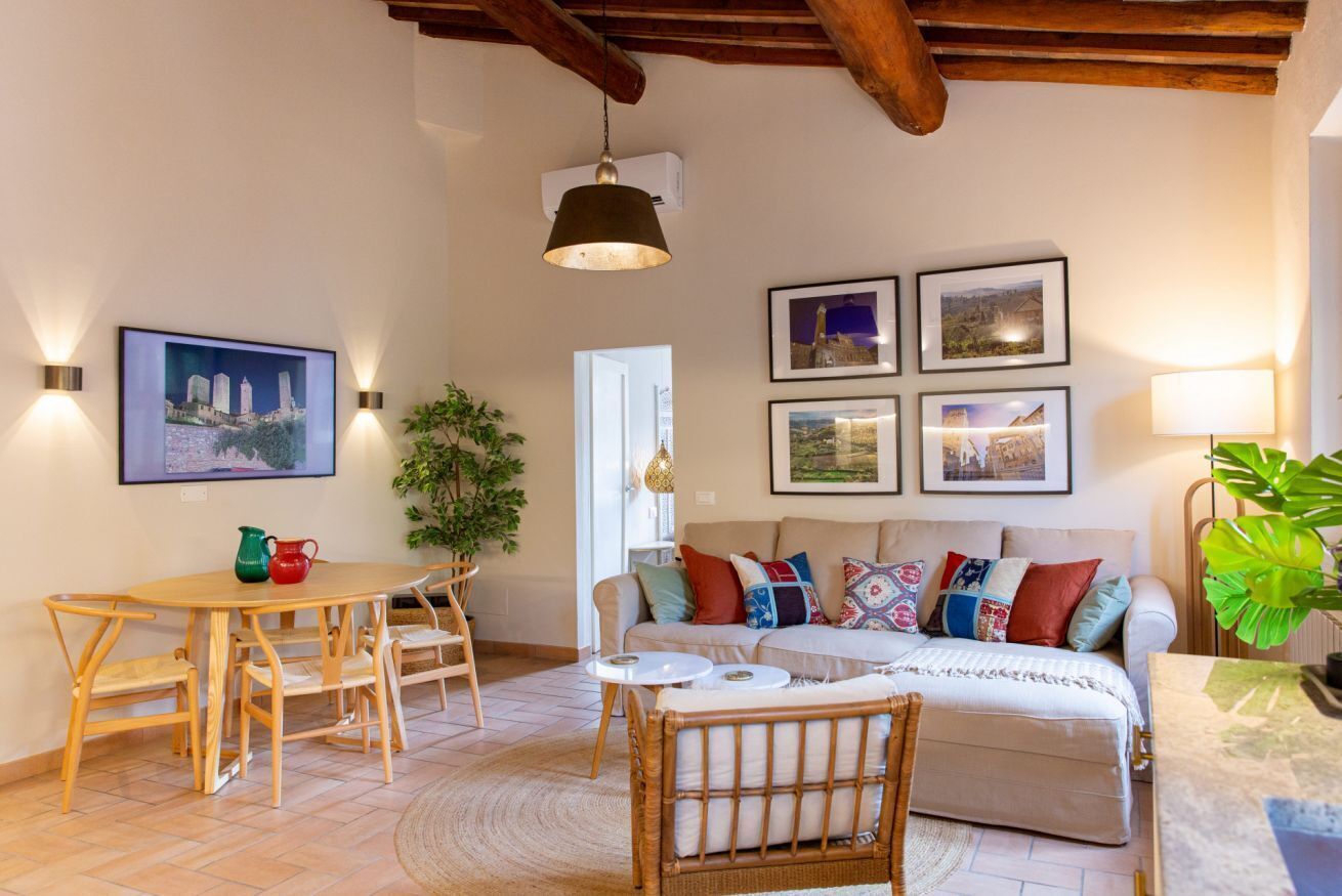 Ein gemütliches Wohnzimmer mit einem Holztisch, Stühlen, einer Couch mit bunten Kissen und gerahmten Bildern an der Wand.