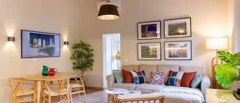 Ein gemütliches Wohnzimmer mit einem Holztisch, Stühlen, einer Couch mit bunten Kissen und gerahmten Bildern an der Wand.