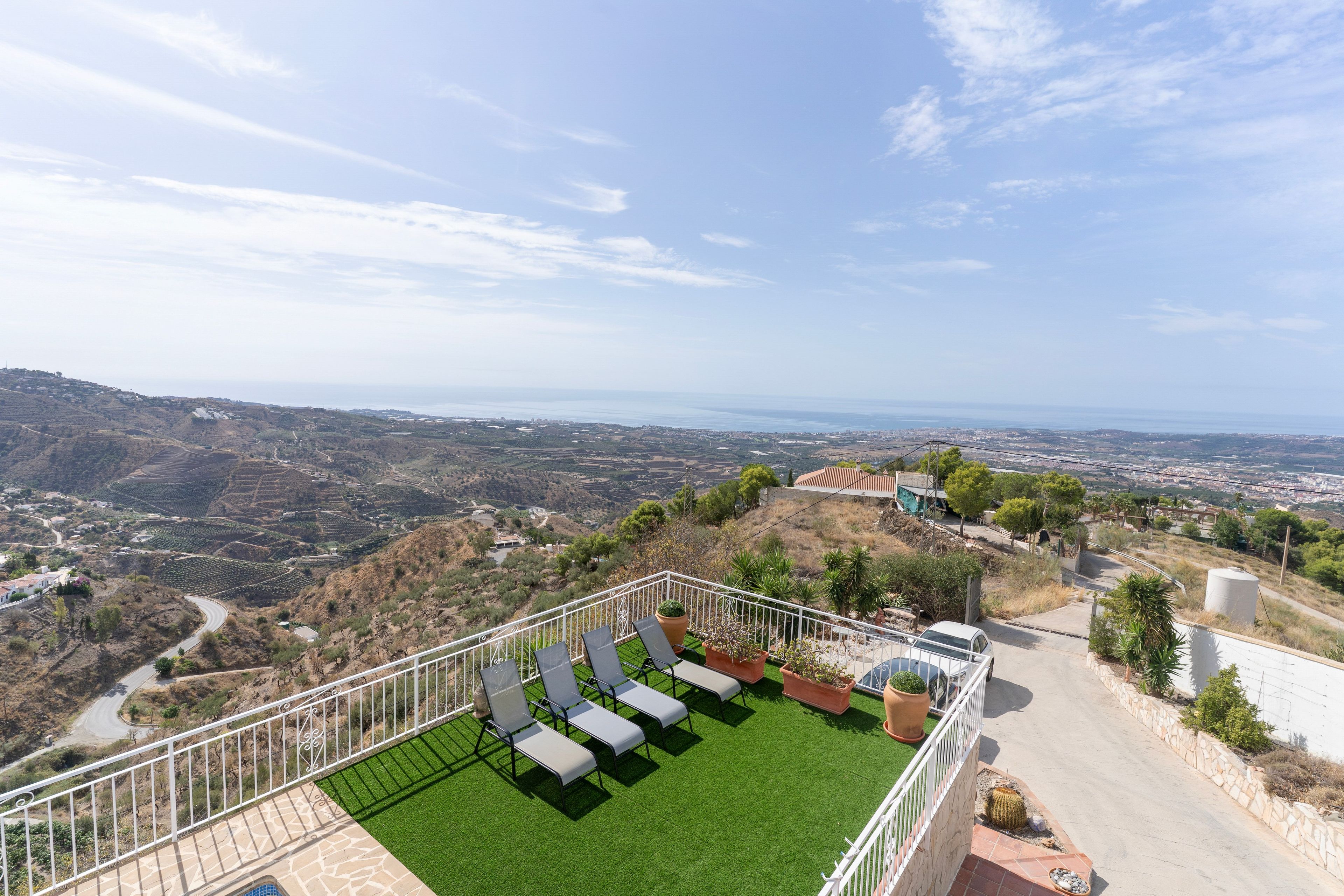 Una terraza con césped artificial y mobiliario exterior, con vistas a un paisaje costero con campos escalonados y una ciudad distante.