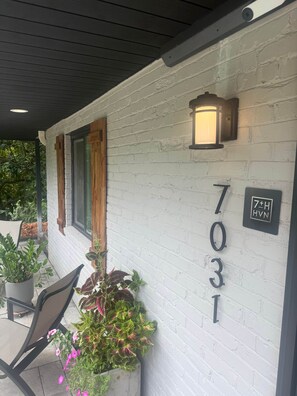 Le numéro de maison, 7031, est peint en blanc sur un mur de briques, et on y voit une plante en pot avec des fleurs roses, ainsi qu’une chaise.
